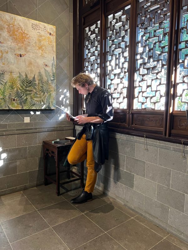 Garrett Bangert leaning on a wall reading a book with sunlight coming through the windows behind him.