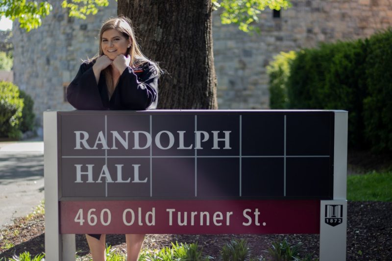 Katelyn Kleinschmidt standing in front of the sign for Randolph Hall on Virginia Tech's campus wearing a regalia.